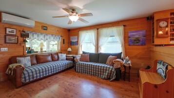 A cozy cabin interior with a wooden floor, a ceiling fan, a sofa, a small table, and a clock on the wall.