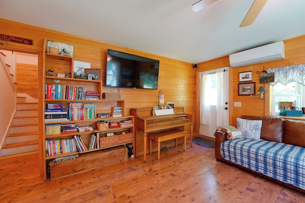 Una habitación acogedora con piso de madera, un piano, una estantería, un televisor y una cama con cubrecolcha estampado.