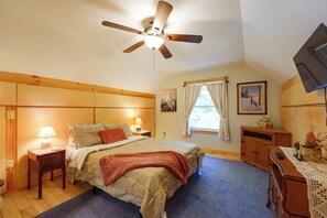 Une chambre à coucher comprenant un lit, des tables de chevet, un ventilateur de plafond, une fenêtre avec des rideaux, un bureau avec une chaise et un tableau accroché au mur.