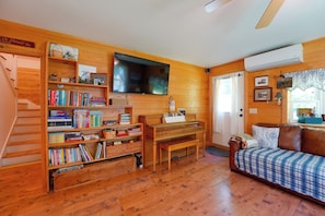 Una habitación acogedora con piso de madera, un piano, una estantería, un televisor y una cama con cubrecolcha estampado.
