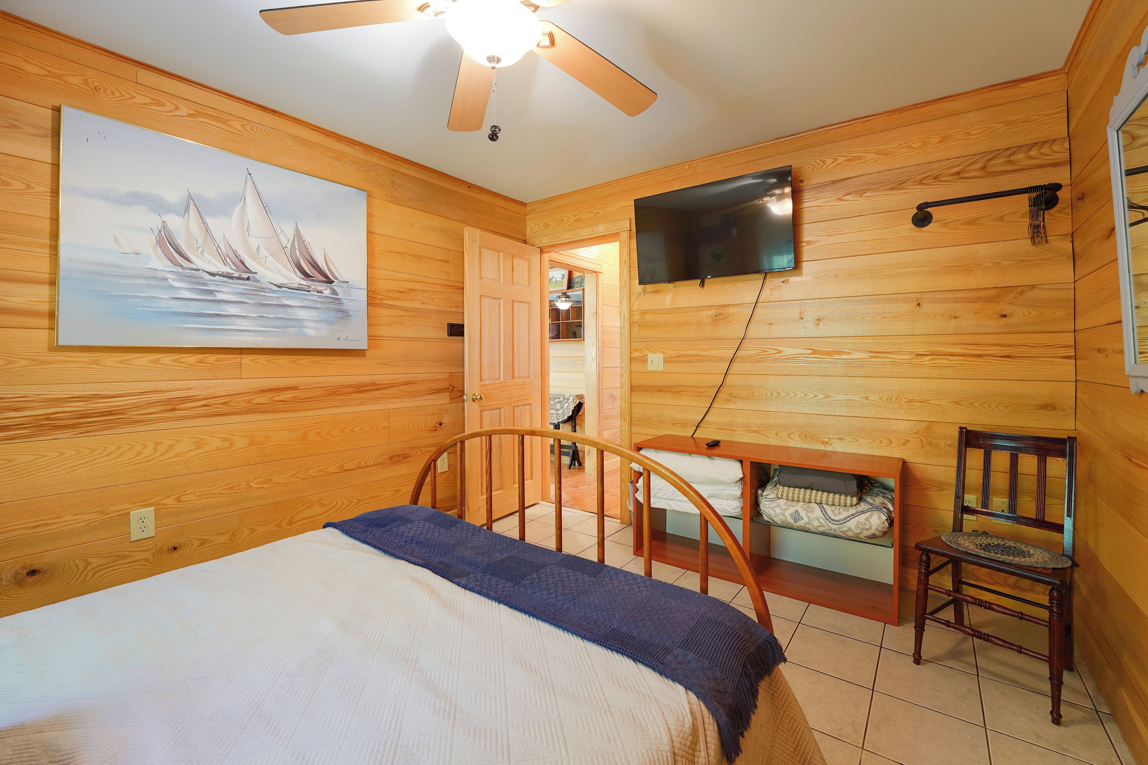 A bedroom with a bed, a ceiling fan, a painting, a television mounted on the wall, and a wooden chair.