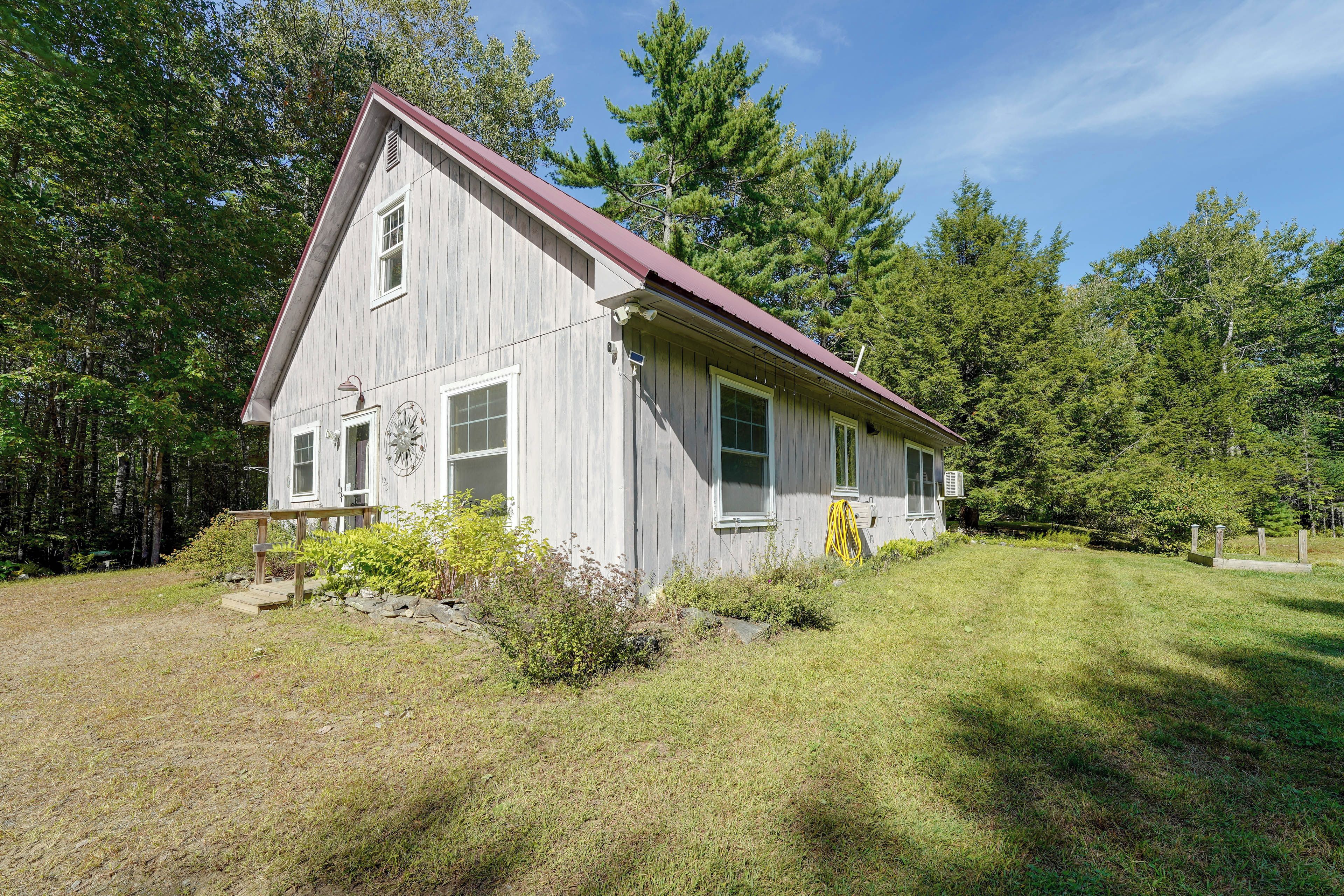 A single-story wooden house with a red roof, surrounded by a grassy yard and dense trees.