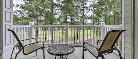 A covered patio with two chairs and a table, a white railing, and a view of trees and a lawn.