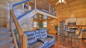 A wooden cabin interior with a leather sofa, dining table, chairs, kitchen area, and a staircase with a balcony. - Gatlinburg Cabin w/ Pool Table & Hot Tub! (Sevierville)