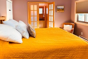 Une chambre avec un grand lit, une porte en bois donnant sur une salle de bain et une fenêtre avec des stores.
