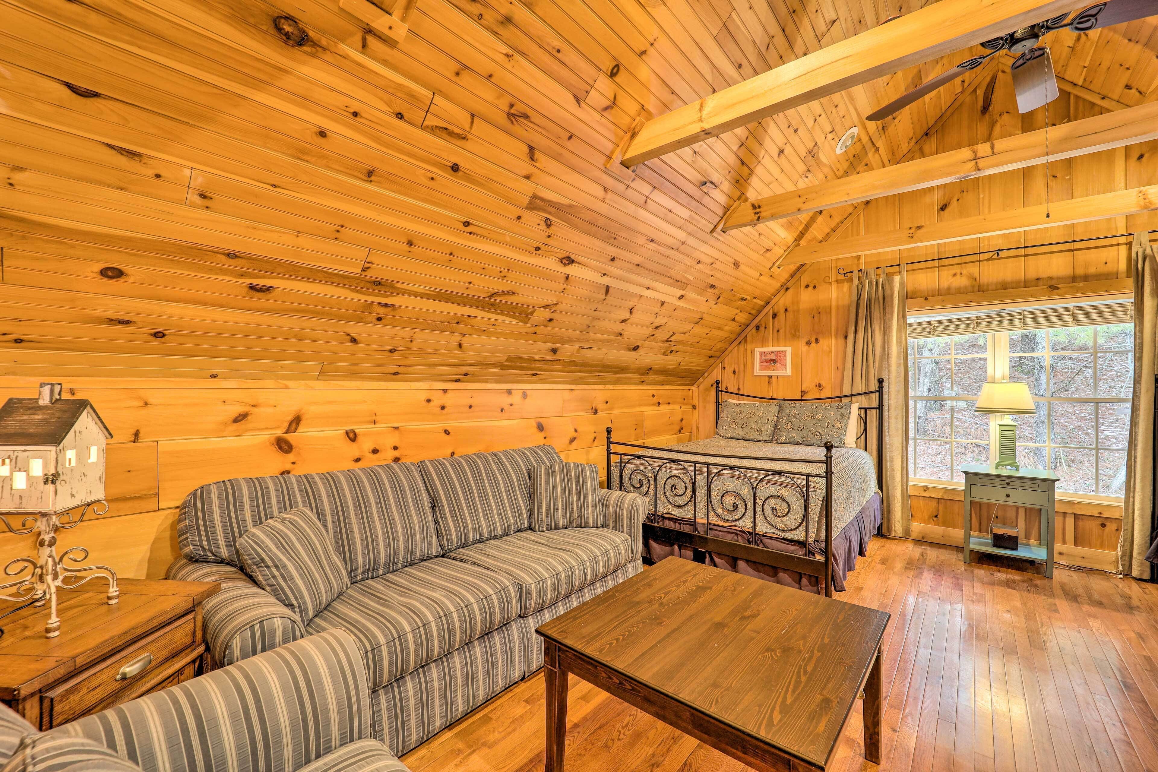 A cozy cabin interior with a wooden ceiling, a bed with a metal frame, a striped sofa, a wooden table, and a window with curtains.