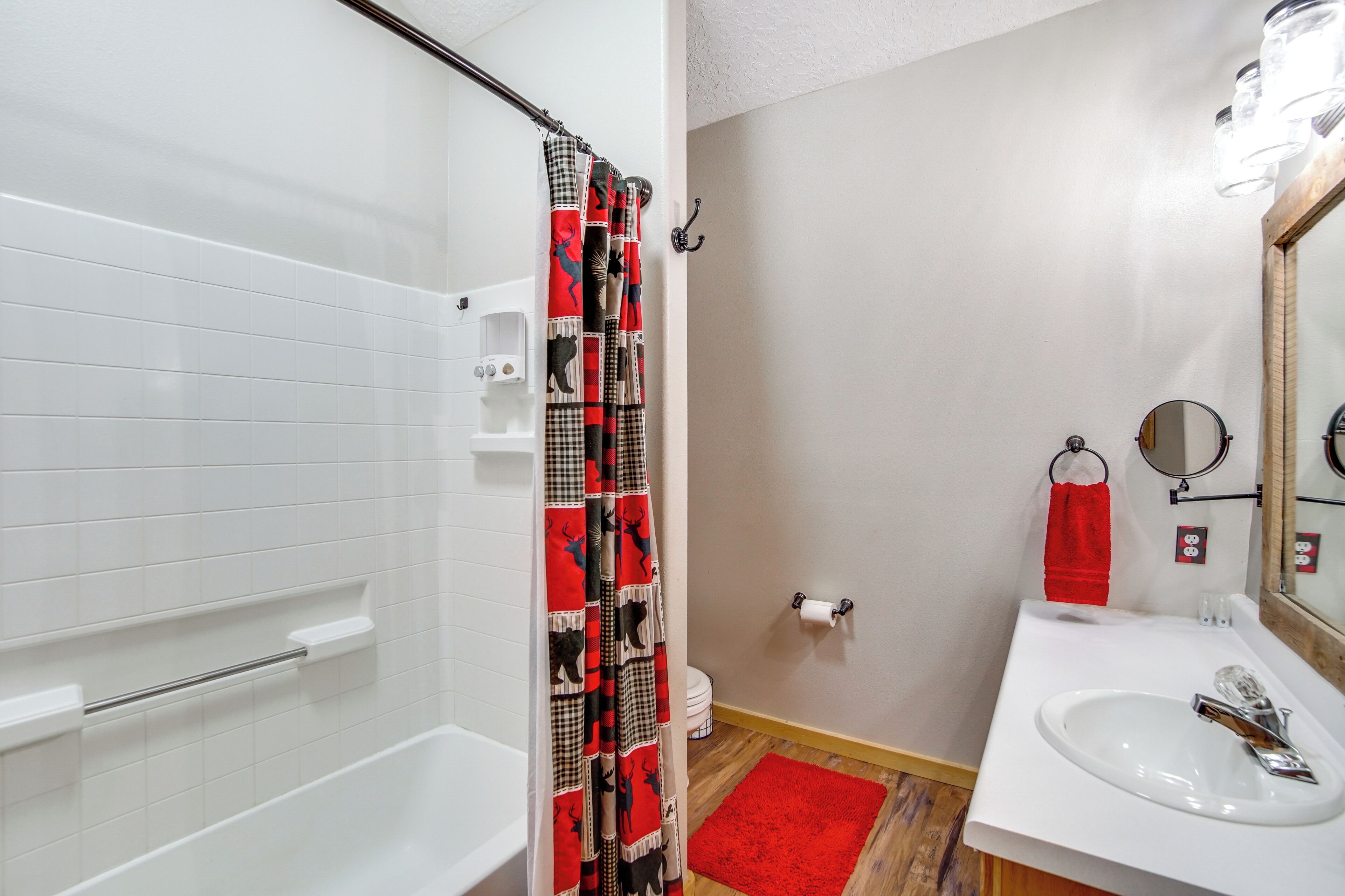 Un baño con una bañera blanca, cortina de ducha con estampado de ciervo, tapete de baño rojo, lavabo blanco y espejo con marco de madera.
