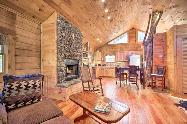 A cozy cabin interior with a stone fireplace, wooden furniture, and a kitchen area.