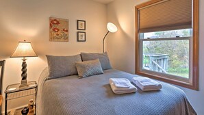 Ein ordentlich bezogenes Bett mit grauer Decke und Kissen, eine Nachttischlampe, ein Fenster mit Jalousien und Blick auf eine Holzterrasse.