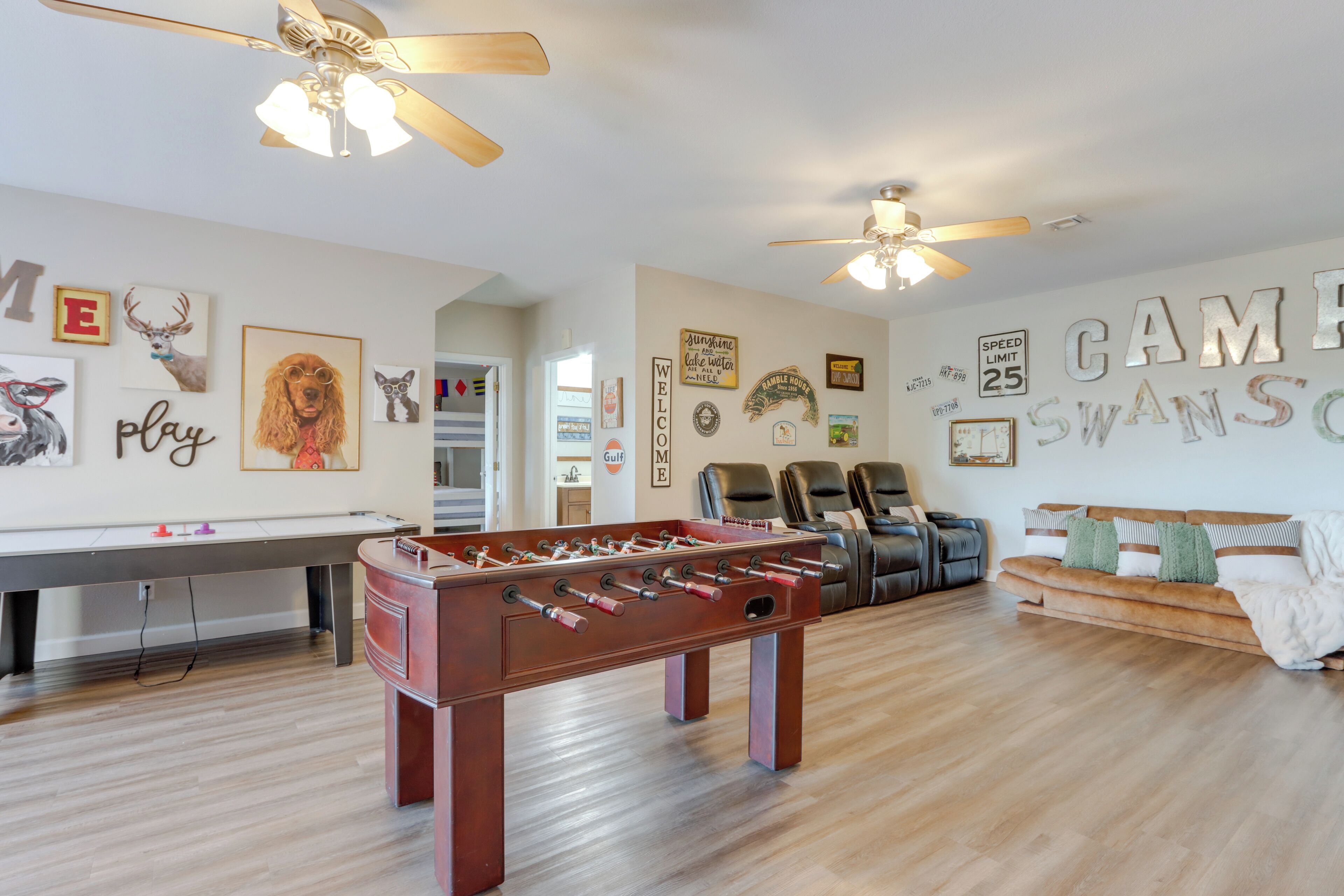 A spacious room with a foosball table, two ceiling fans, a sofa, and a wall with various framed pictures and signs.