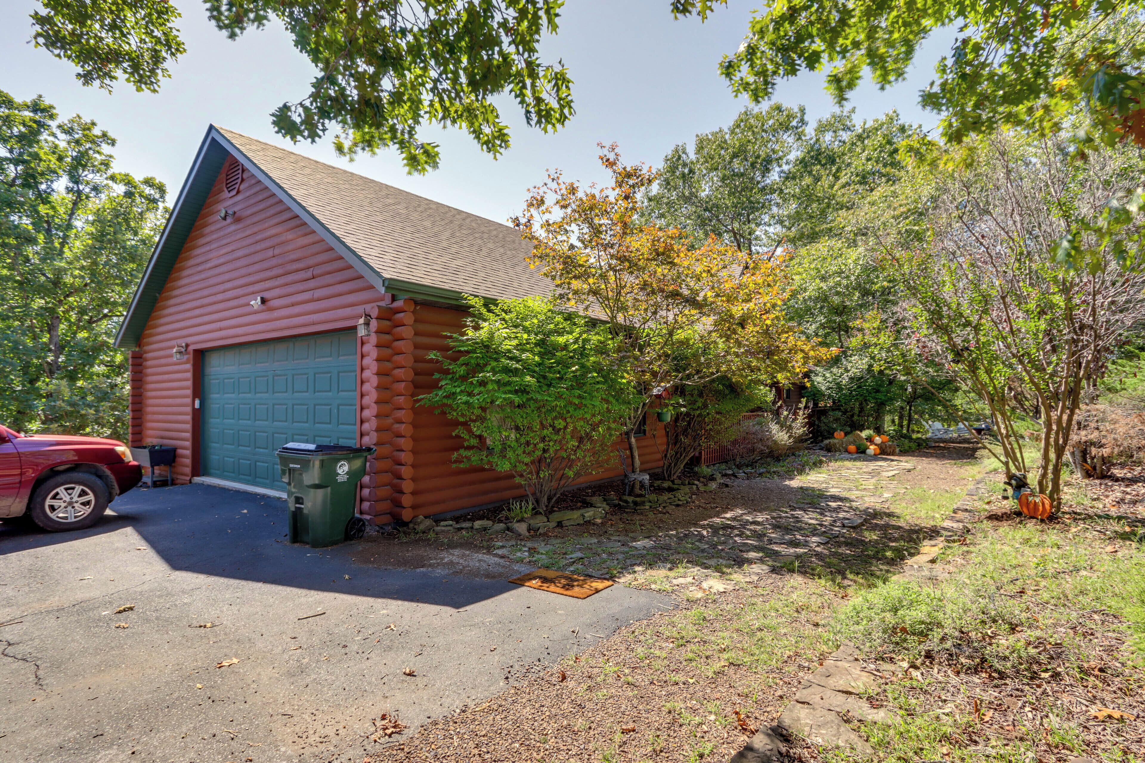 A log cabin with a garage, a red car parked in front, and a green trash bin.