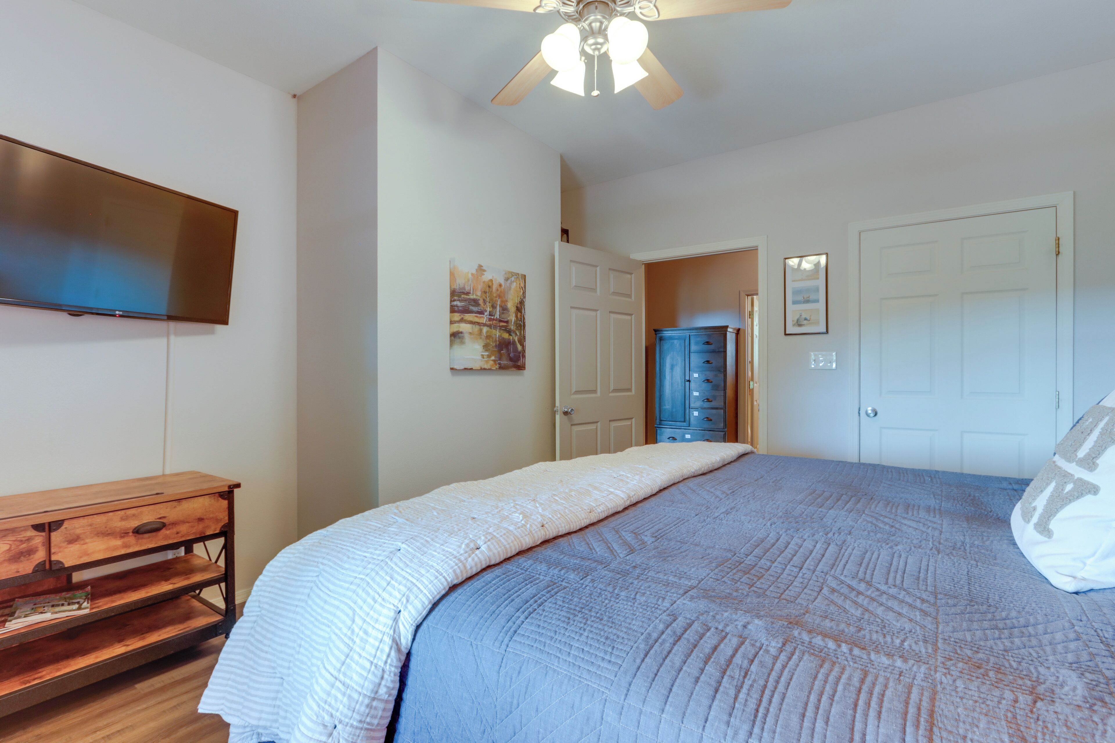 A bedroom with a bed, a ceiling fan, a TV, a dresser, and a door leading to another room.