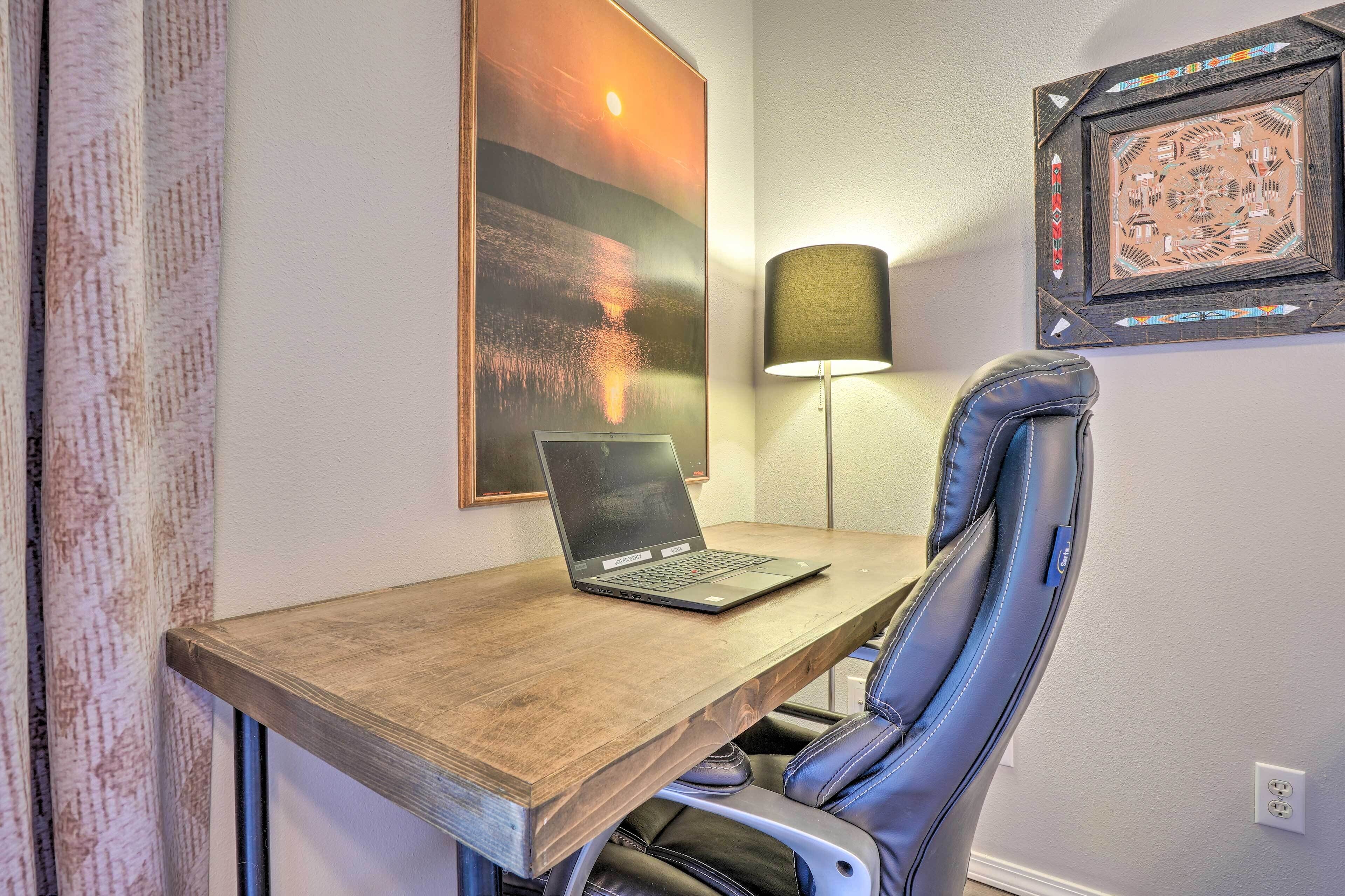 Ein Holztisch mit einem Laptop, ein Ledersessel, eine Wandlampe und ein gerahmtes Kunstwerk.