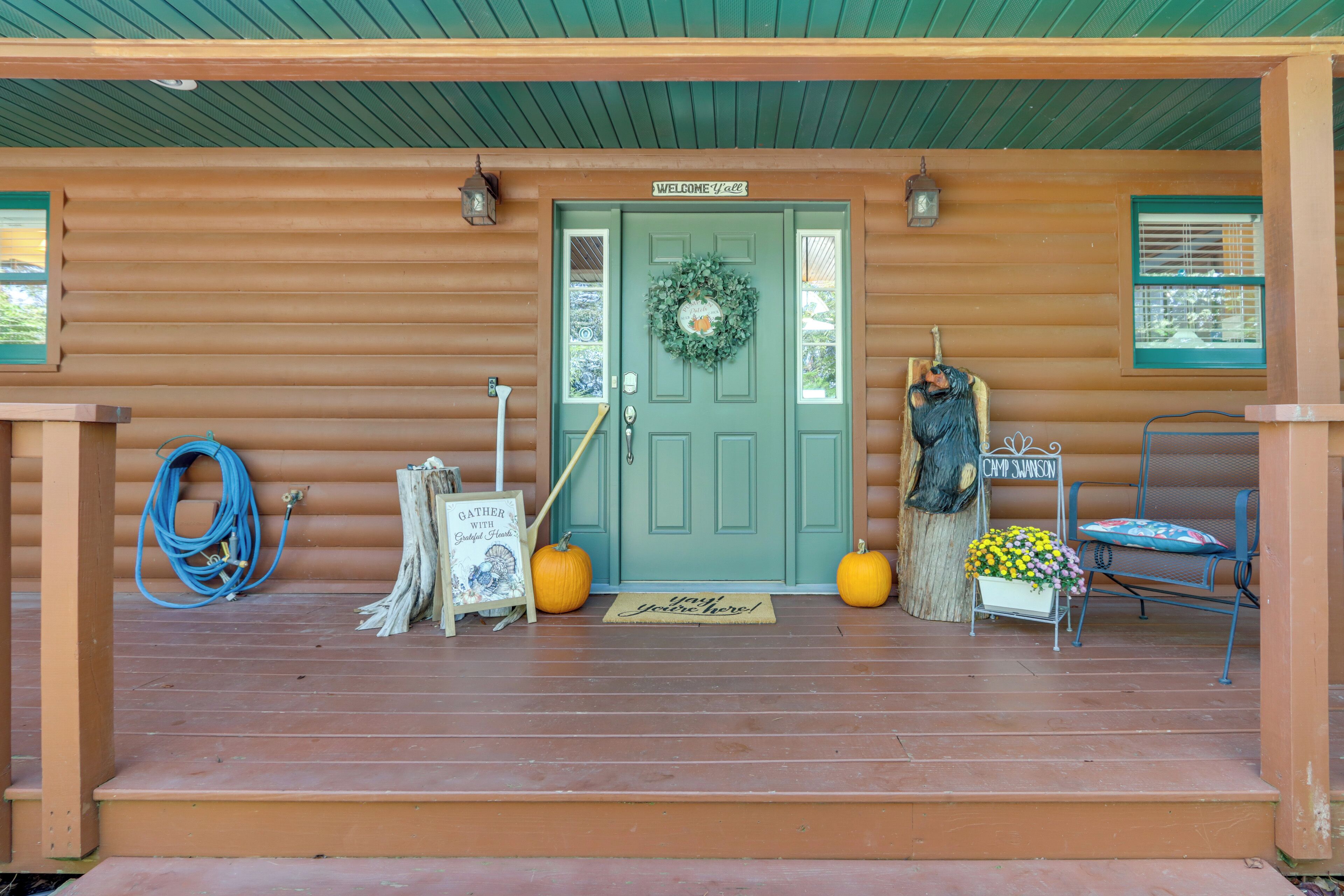 A wooden cabin with a green door, a welcome sign, a wreath, a sign reading "Camp Samson," two pumpkins, a chair, and a sign that says "Gather with Friends."