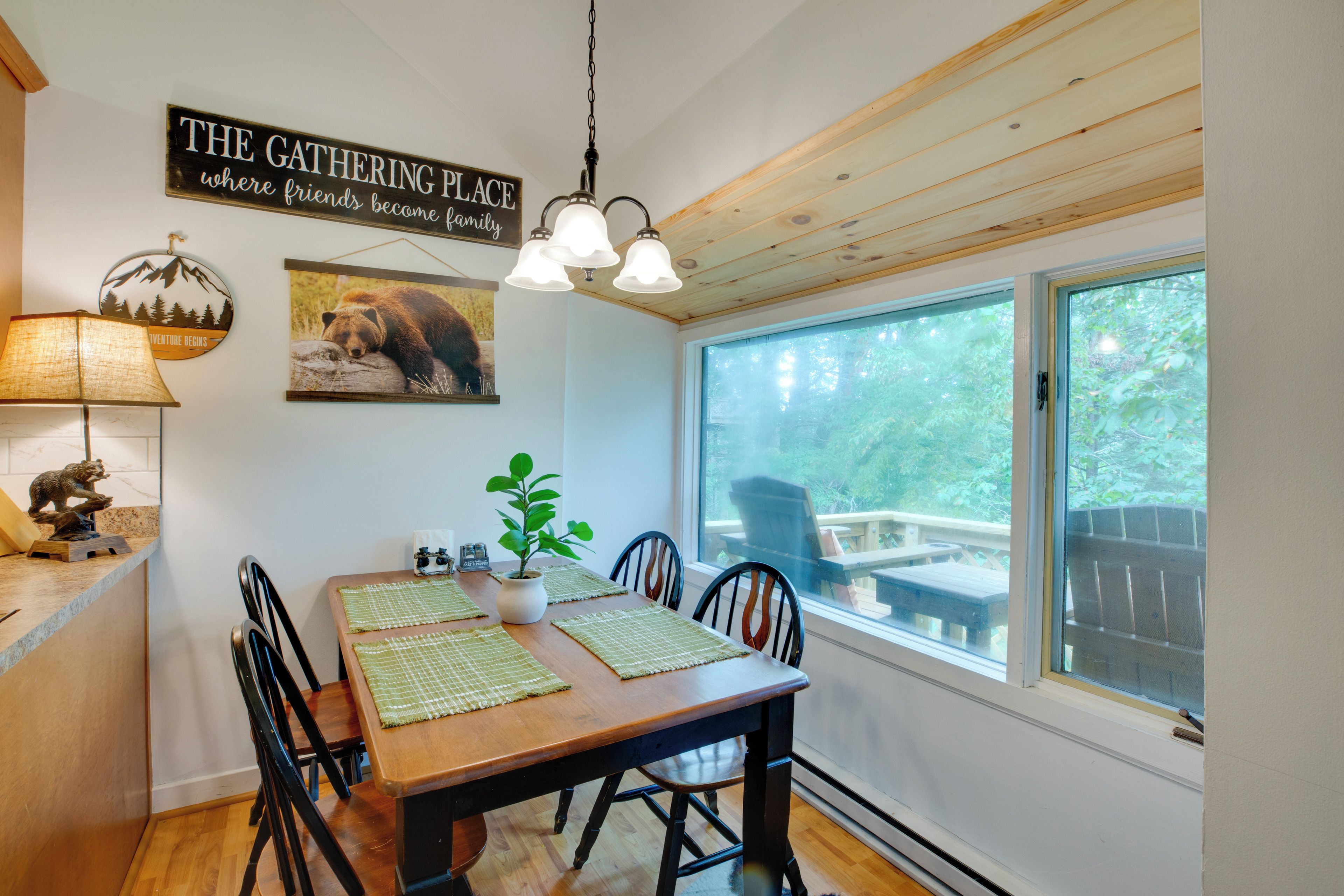 Een eetruimte met een houten tafel, stoelen, een hanglamp, een wandbord en een raam met uitzicht op de omgeving.