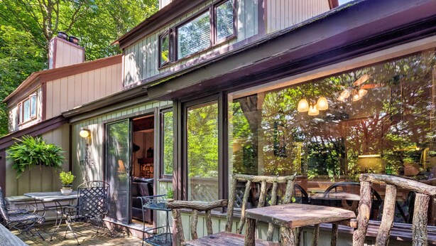 A wooden deck with rustic furniture, large windows, and a view of the surrounding greenery.