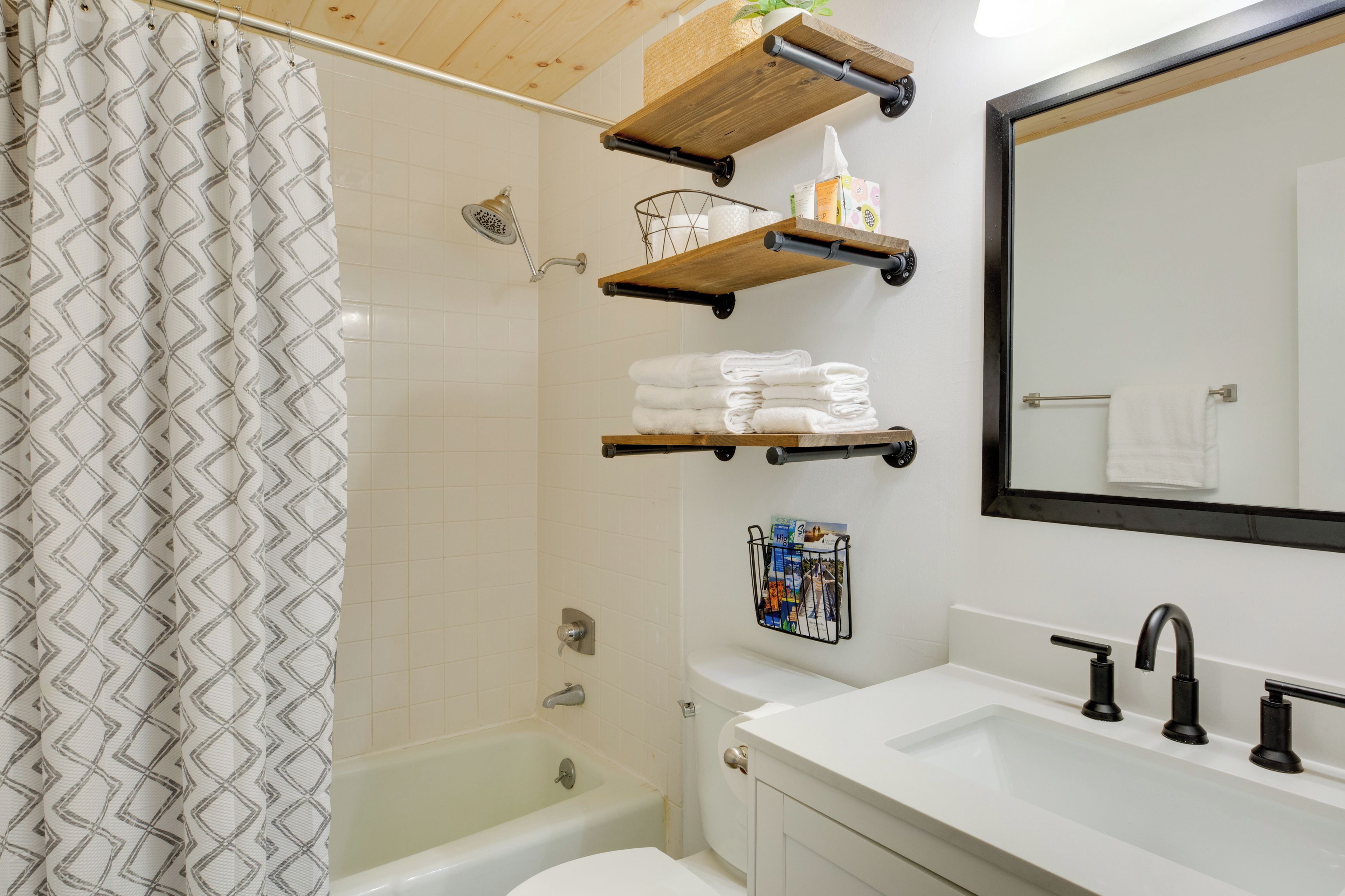 A bathroom with a shower, a toilet, a sink, and a mirror. There are towels stacked on a shelf, and a magazine rack with a magazine.