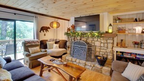 A cozy cabin interior with a stone fireplace, wooden furniture, and a large window overlooking greenery.