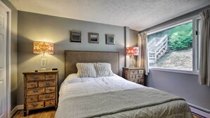A bedroom with a large bed, two nightstands, a dresser, and a window with a view of a staircase and greenery.