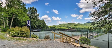 A tennis court with a wooden fence and a bench, surrounded by trees and hills.