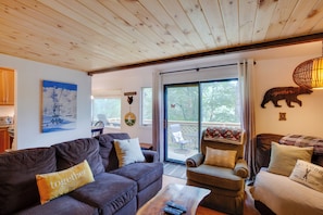 A cozy living room with a wooden ceiling, a sofa set, a coffee table with remotes, and a view through the glass door. - Steps to Sugar Mountain! Condo w/ Furnished Deck (Banner Elk)