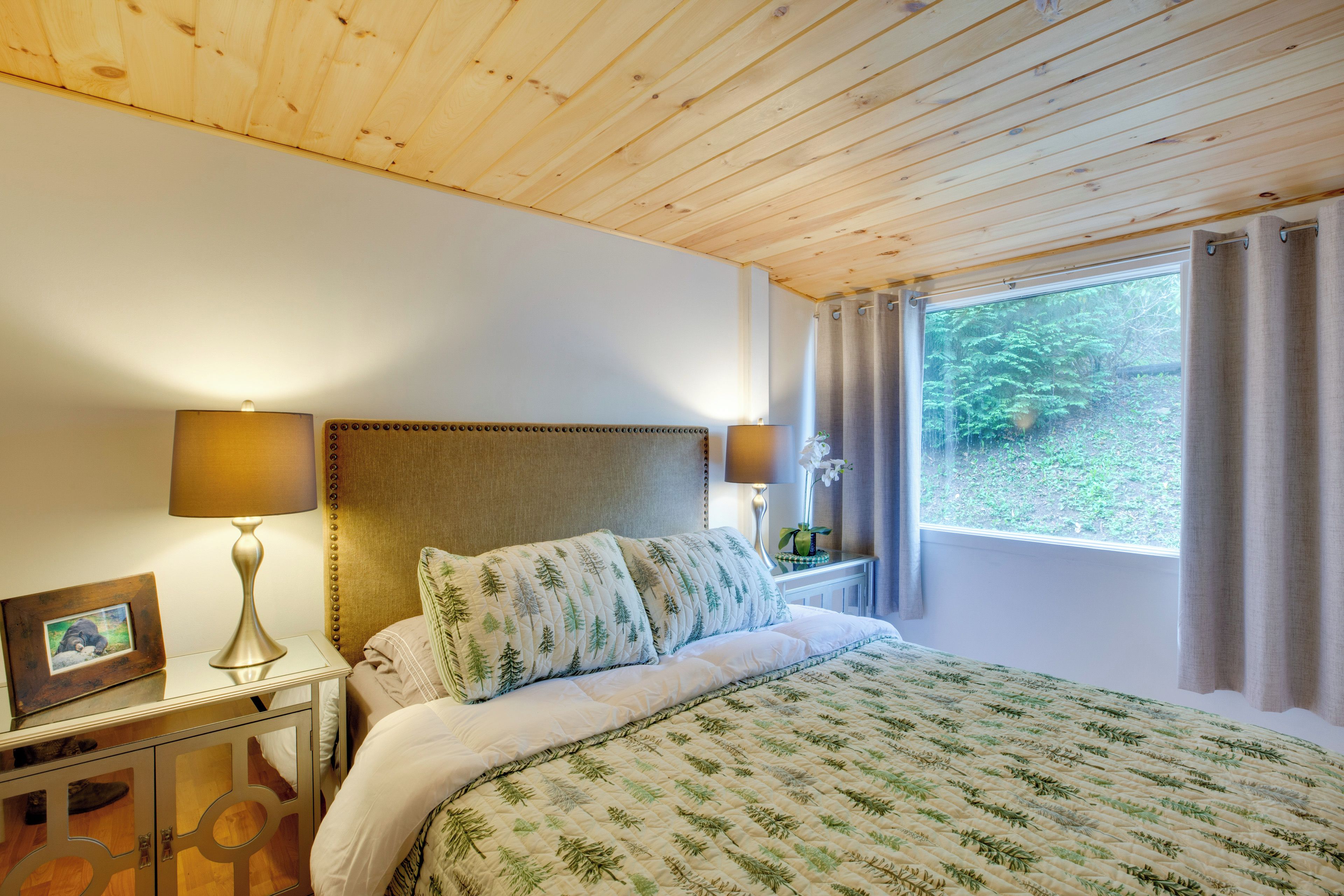 A bedroom with a wooden ceiling, a bed with a green leaf-patterned duvet, two bedside lamps, a nightstand with a framed picture, and a window with curtains.