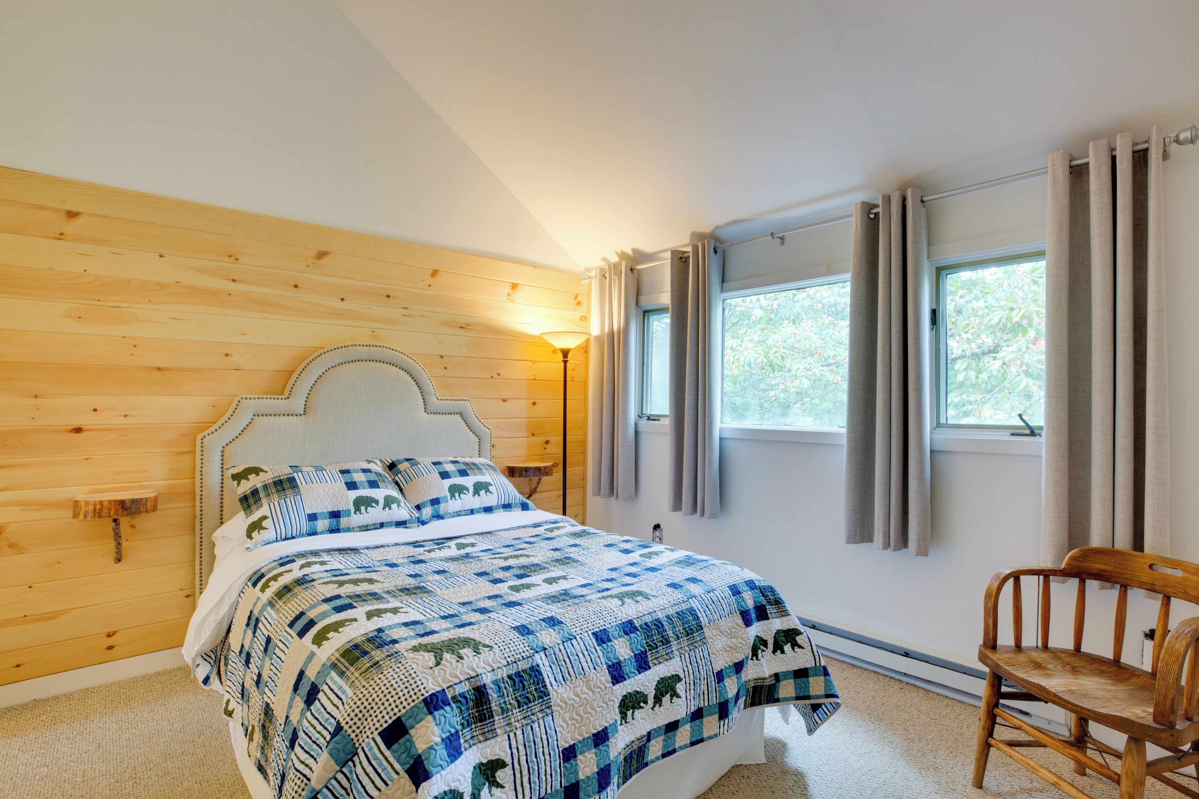 Ein Schlafzimmer mit einem hölzernen Kopfteil, einer gesteppten Bettdecke, einem Nachttisch, einem Sessel und einem Fenster mit Vorhängen.