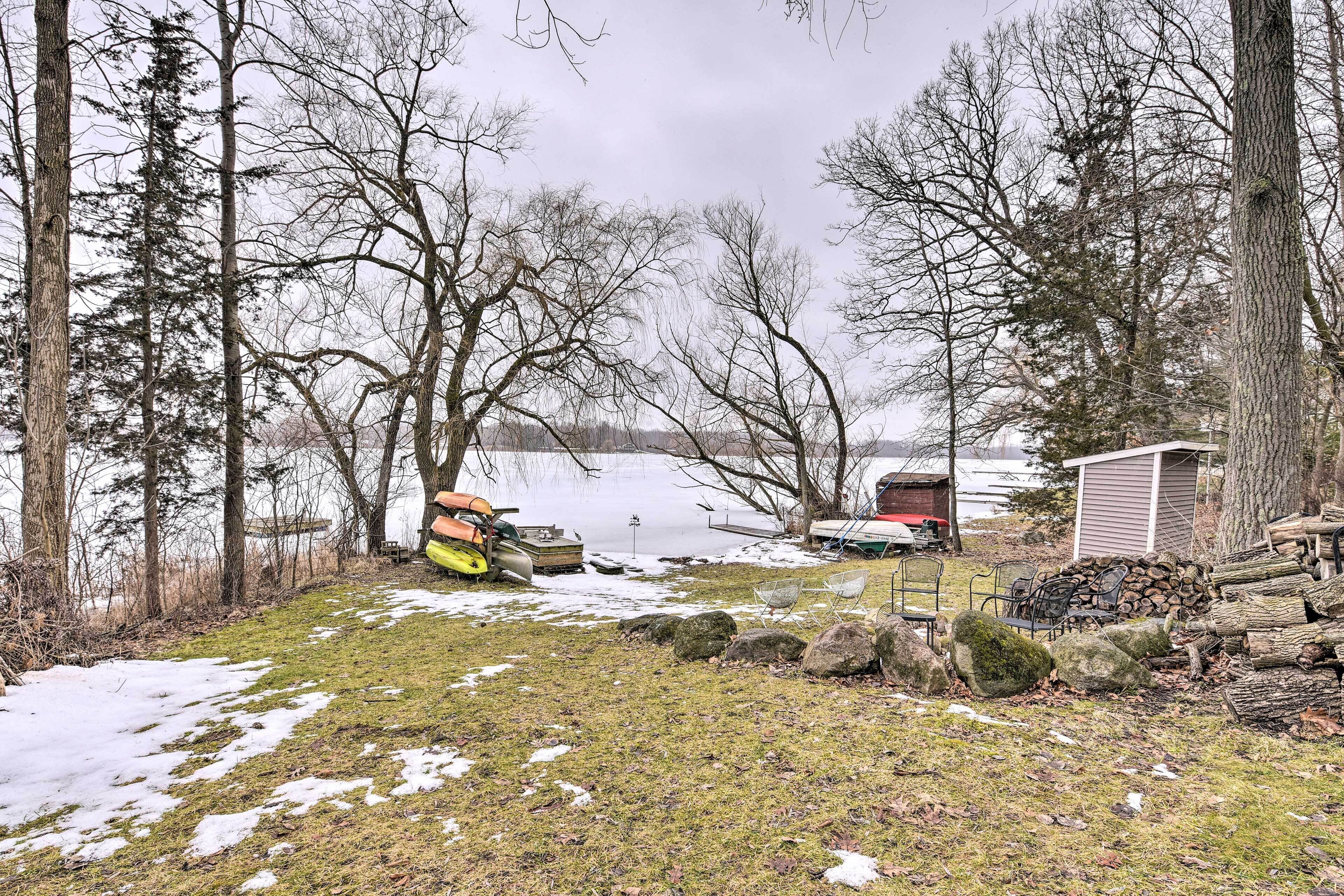 A lakeside cabin with a small shed, a kayak, and a pile of firewood.