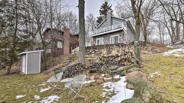 A stone house with a balcony, surrounded by trees and a yard with chairs and a shed.