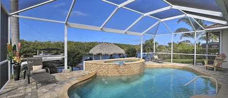 A covered outdoor swimming pool with a hot tub, lounge chairs, and a thatched-roof structure.