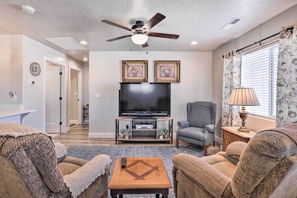 A living room with a ceiling fan, a flat-screen TV on a wooden stand, two framed pictures on the wall, a grey armchair, a beige sofa, and a lamp on a wooden side table.