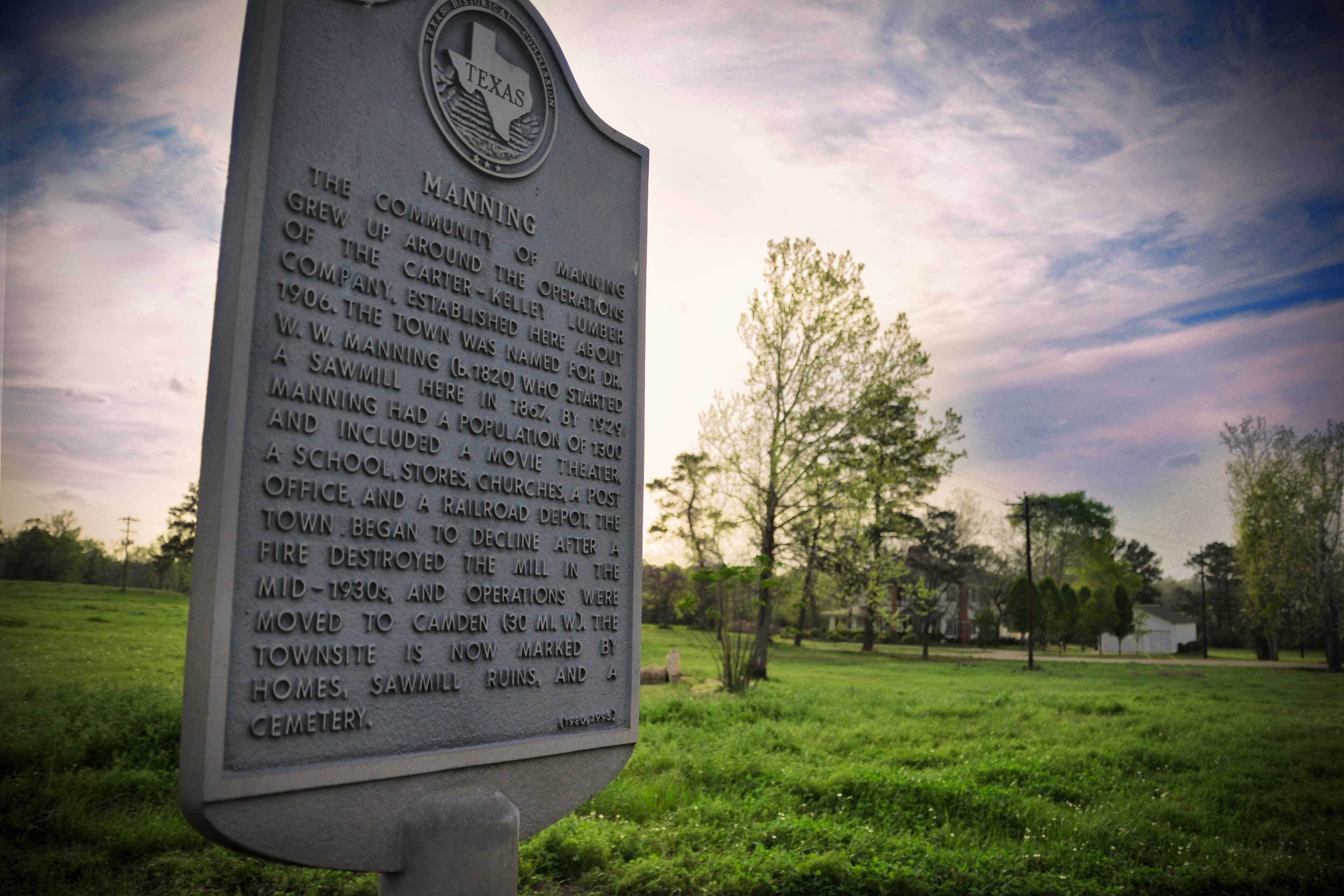 A historical marker in a grassy area with trees and a cloudy sky in the background.