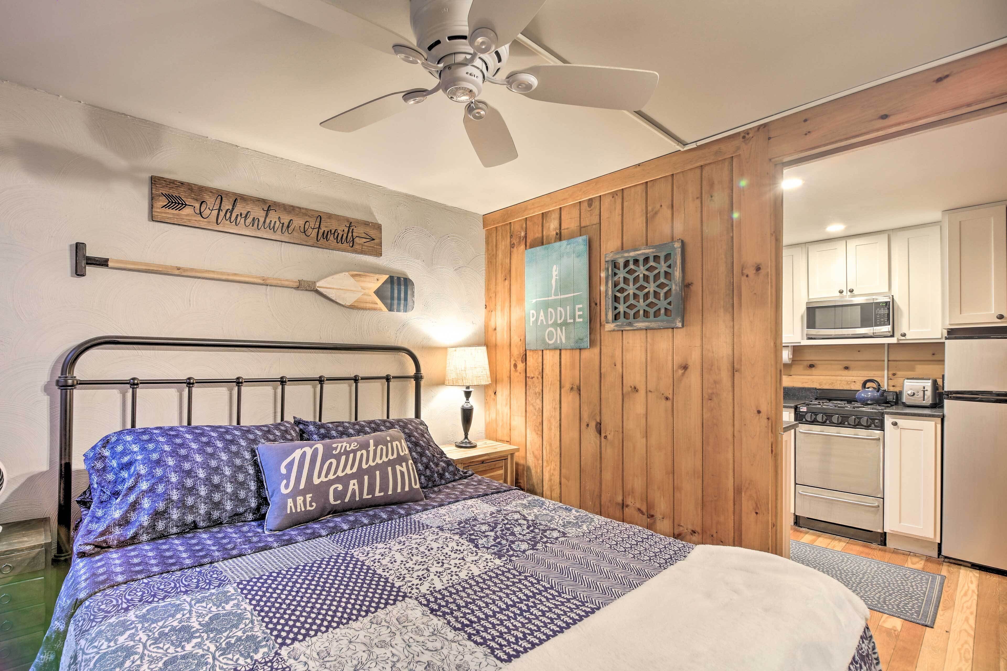 Ein Schlafzimmer mit Bett, Holzwand, Deckenventilator, Nachttisch, Lampe und einer Küchenzeile mit Schränken und Geräten.