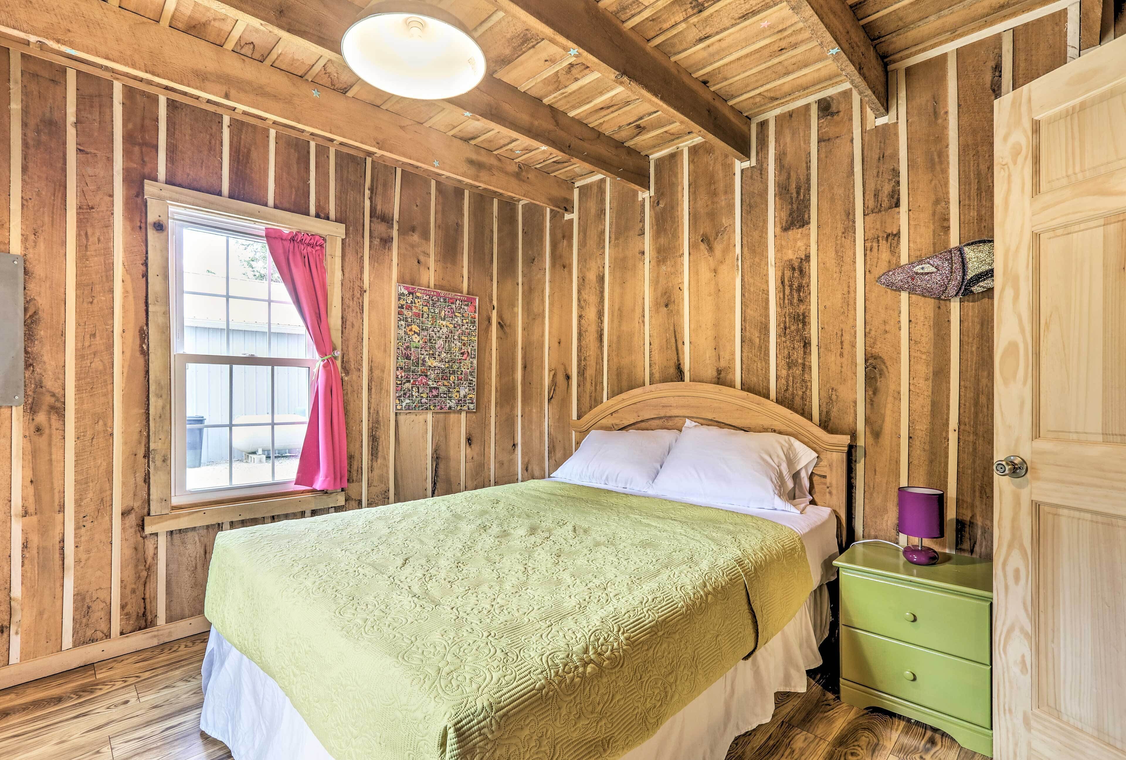 Une pièce en bois avec un lit, une fenêtre ornée de rideaux roses, une table de chevet verte et un plafond en bois.