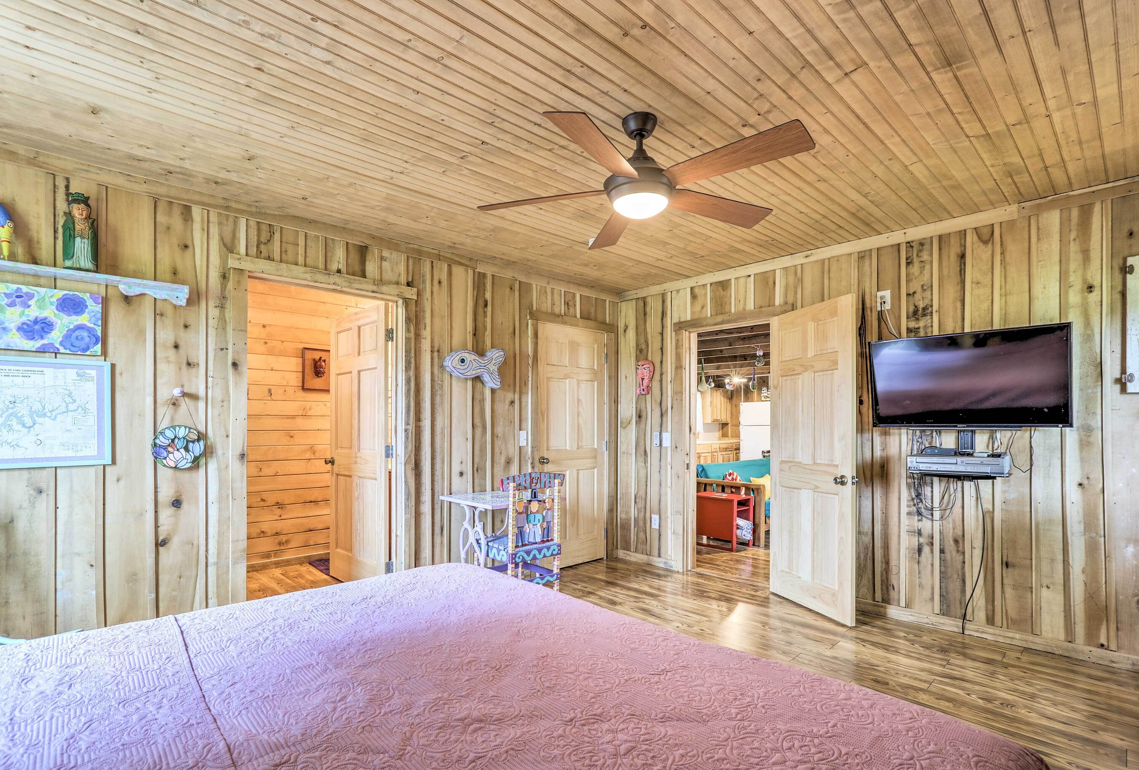 Une pièce aux murs en bois, avec un ventilateur de plafond, un lit, une télévision et un coin cuisine visible à travers une porte ouverte.