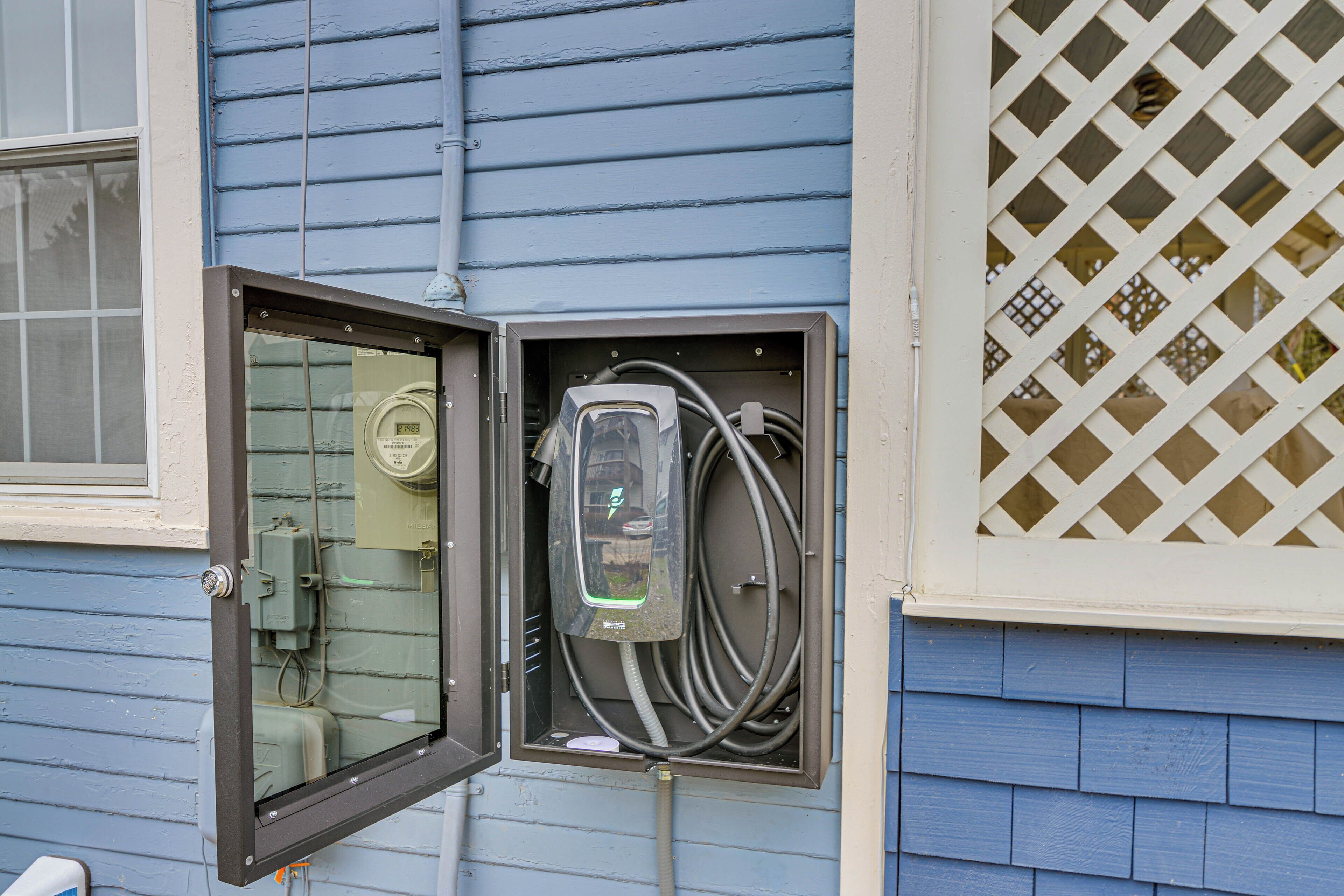 An open electrical box with visible wiring and a meter, attached to a blue house with a window and a lattice window.