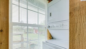 A laundry room with a stacked washer and dryer, wooden walls, and a window with blinds.