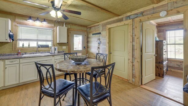 A rustic kitchen with wooden cabinets, a dining table with chairs, a ceiling fan, and a window with blinds.