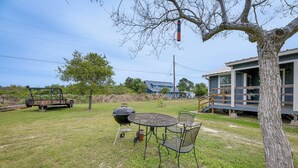 A backyard with a grill, a table, and chairs, a tree, and a house in the background.