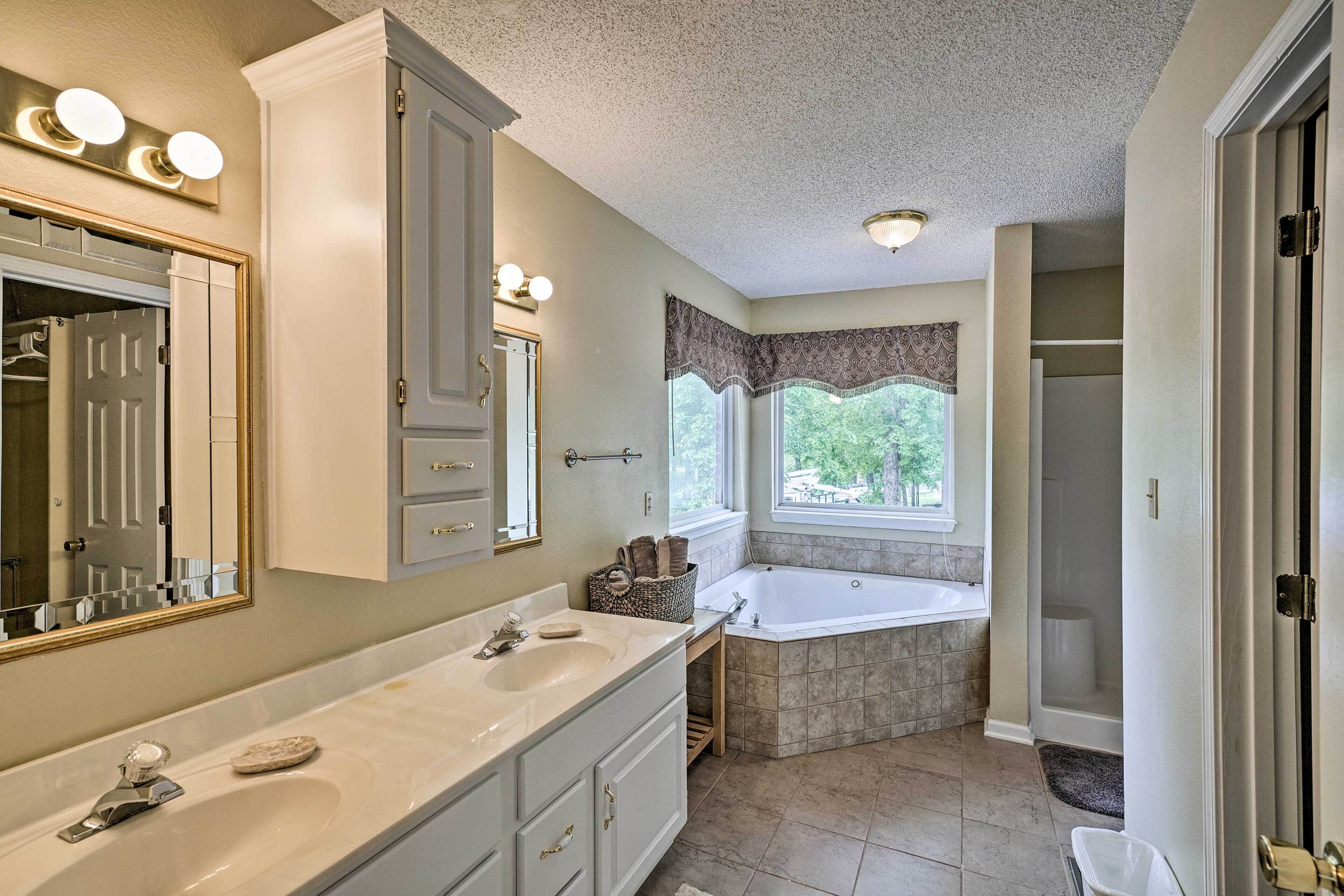 A bathroom with a double sink vanity, a large mirror, a bathtub, and a window with a view of trees.