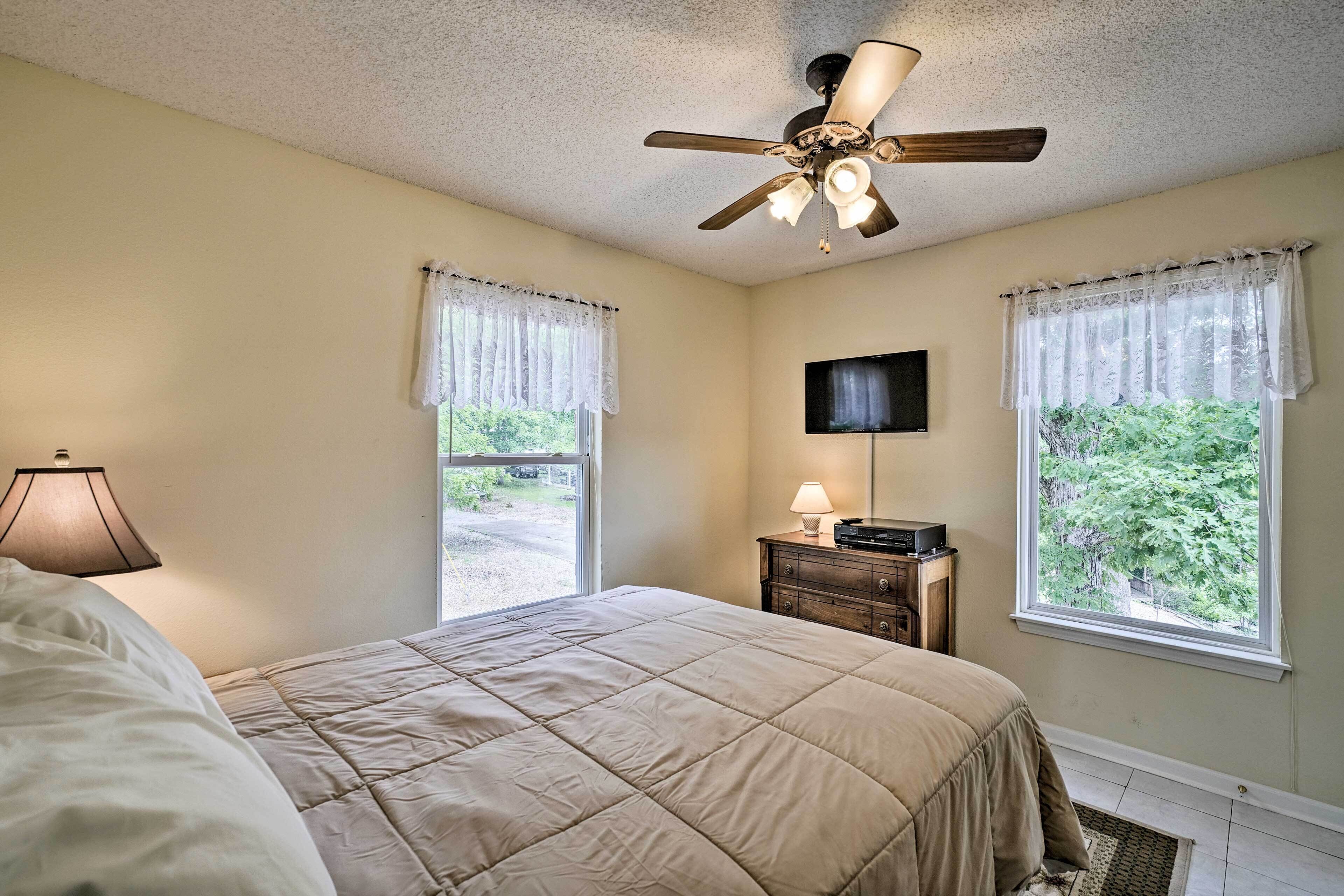 A bedroom with a bed, a ceiling fan, a television, and two windows with curtains.