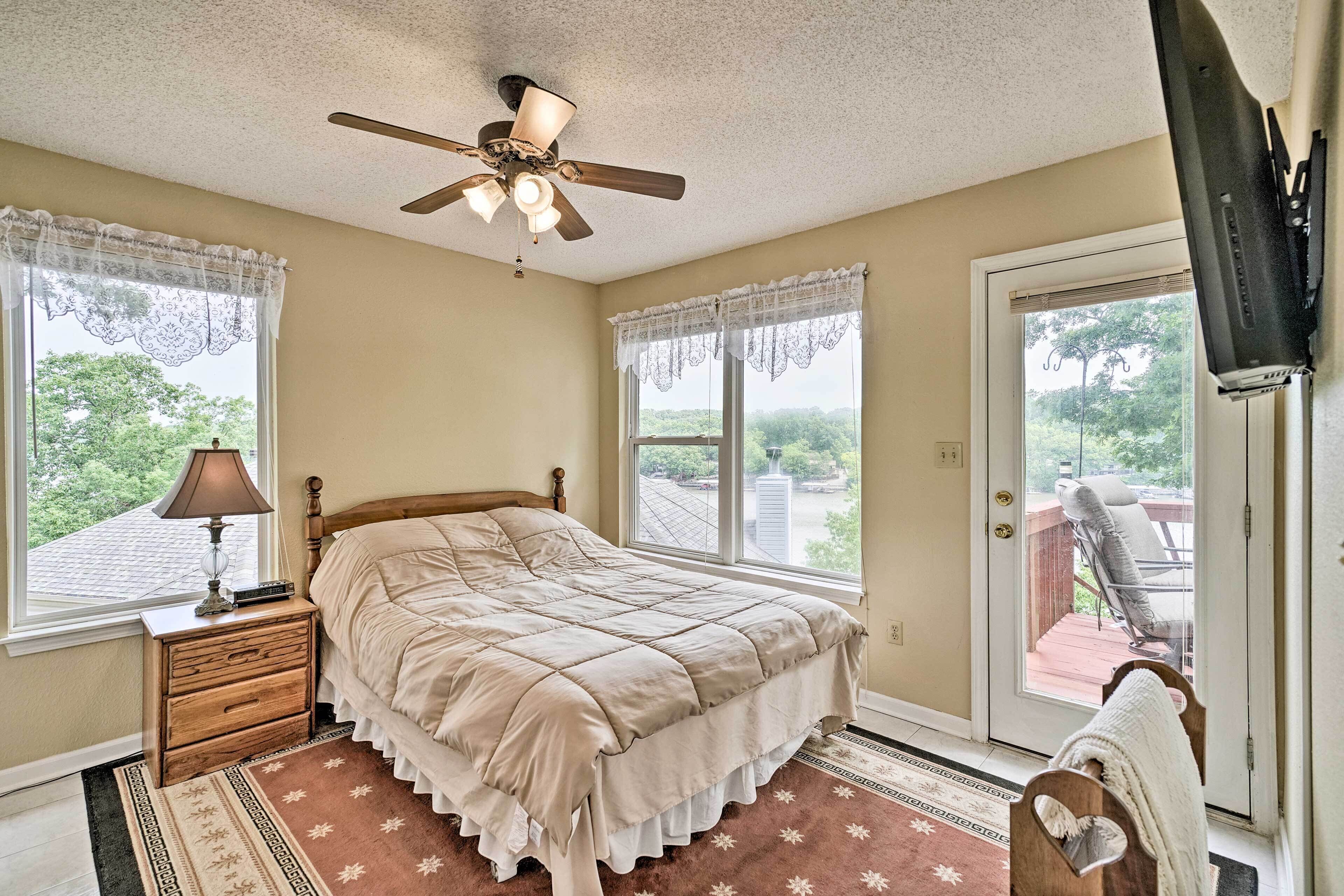 A bedroom with a bed, a ceiling fan, a window with a view, a nightstand, and a television mounted on the wall.