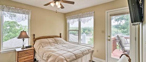 A bedroom with a bed, a ceiling fan, a window with a view, a nightstand, and a television mounted on the wall.