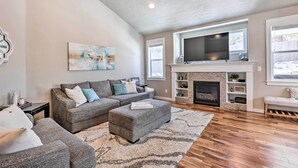 A living room with a grey sectional sofa, a grey ottoman, a fireplace, and a flat-screen TV above it. - Modern Bend Home ~ 3 Mi to Old Mill District! (Bend)