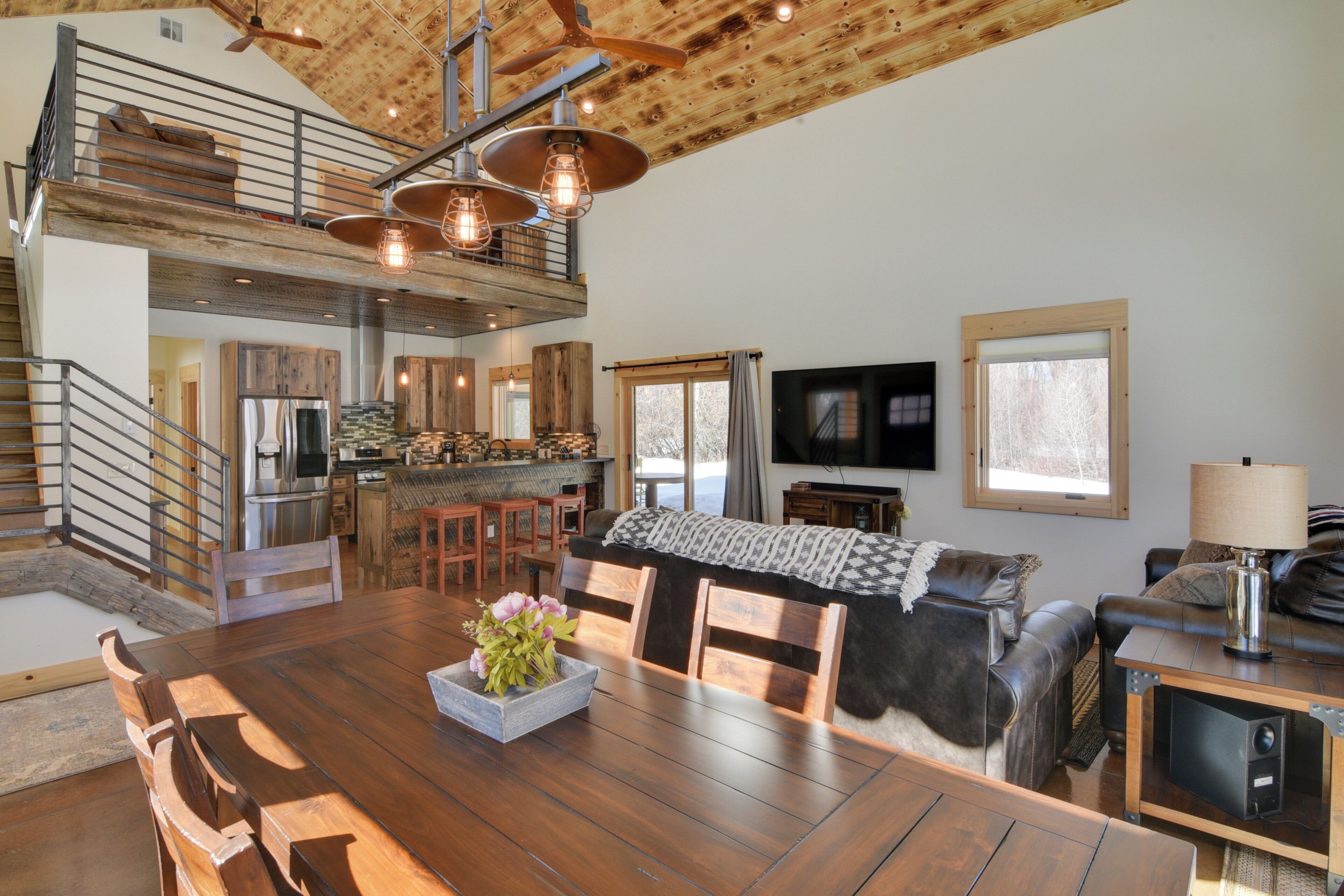 A spacious living area with a wooden dining table, a sofa, and a TV. There is a kitchen area with modern appliances and a staircase leading to an upper level.
