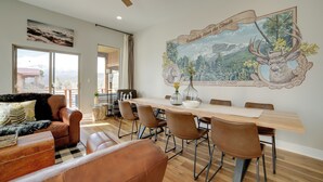 A dining area with a wooden table and brown chairs, a large wall mural, and a view through the window. - Woodland Park Escape: Licensed, Hot Tub, Mtn Views (Woodland Park)
