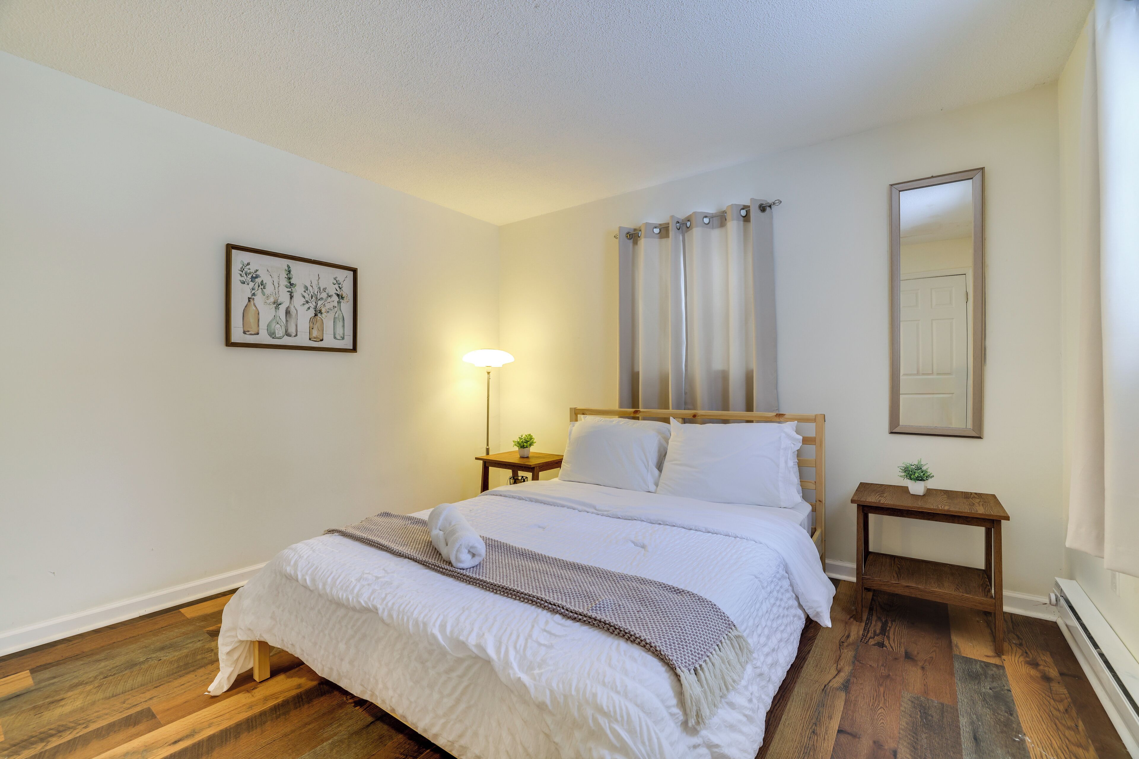 A bedroom with a bed, a bedside table, a lamp, a mirror, and a painting on the wall.