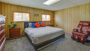 Quarto com cama, paredes de madeira, duas janelas, mesa de cabeceira, cômoda, cadeira de massagem e ventilador de teto.