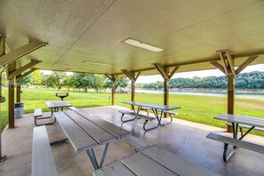 Un espace pique-nique abrité, équipé de plusieurs tables et bancs, offrant une vue sur une zone herbeuse et une étendue d’eau.