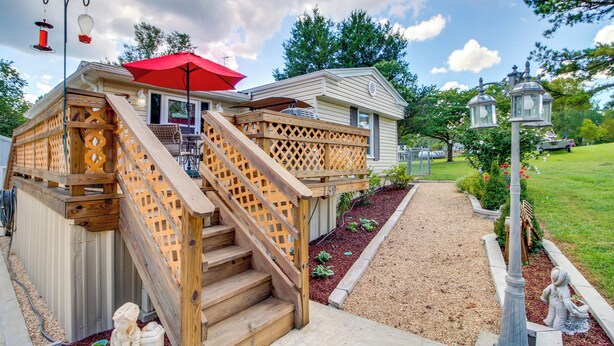 A wooden deck with a staircase leading to a covered area, a small building with a red umbrella, and a garden with a statue. - Bull Shoals Lake Home at Beaver Creek Marina! (Kissee Mills)